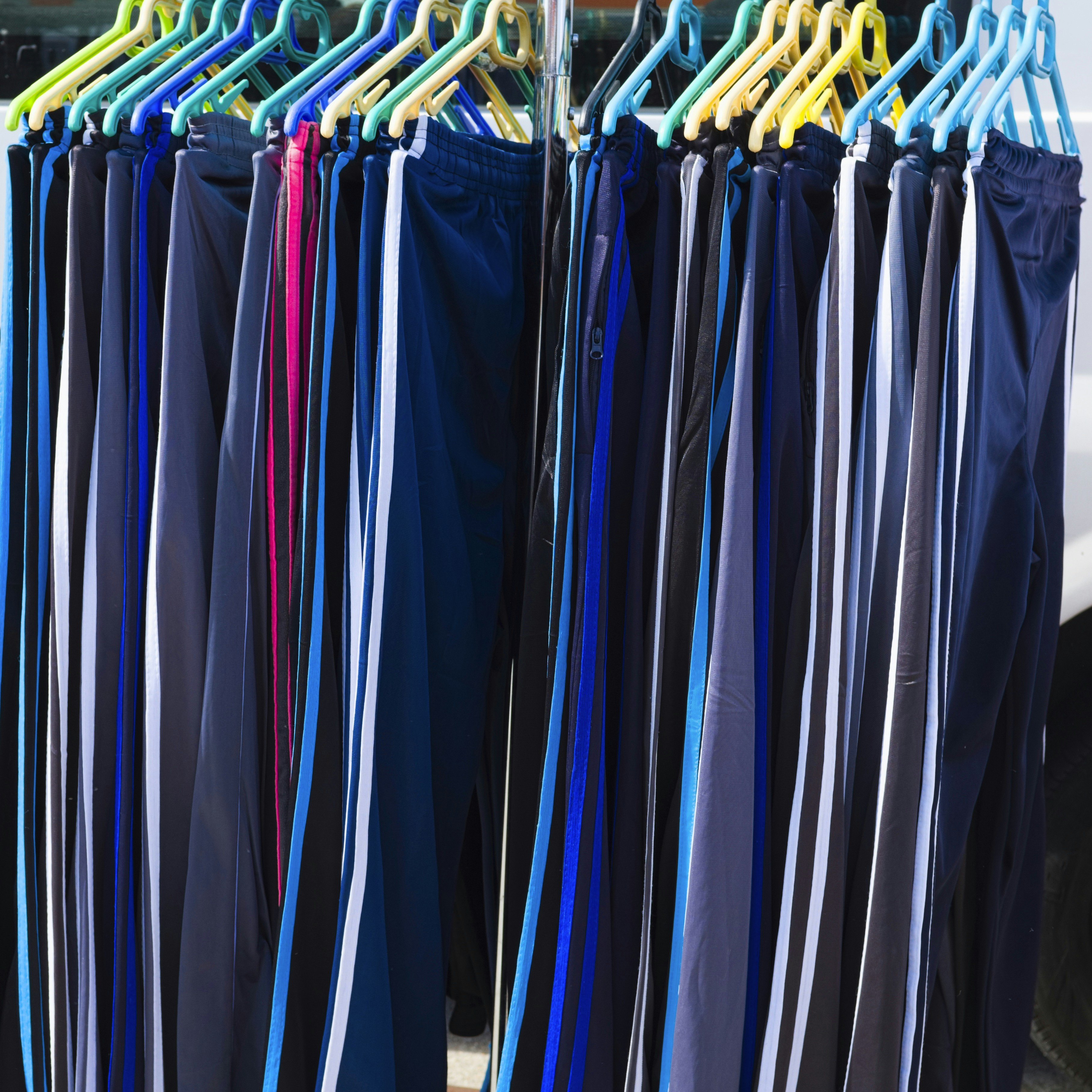 Rows of Blue and Black Sweatpants Hanging on Hangers in a Market - A rack of blue and black sweatpants with white stripes hang on plastic hangers, likely in an outdoor market setting. The pants are all neatly arranged and show a variety of sizes and styles.